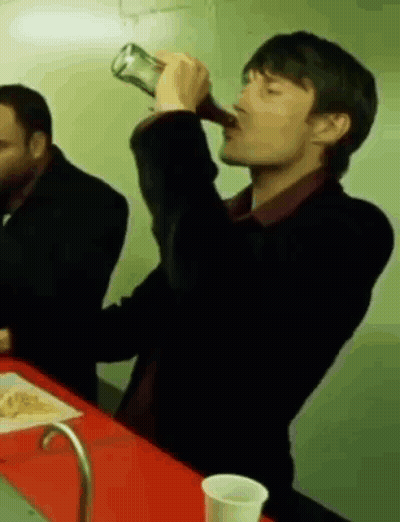 A Man Is Drinking From A Bottle While Sitting At A Table With Another Man . GIF