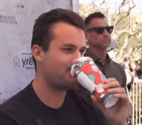 A Man Is Drinking From A Red And White Cup While Another Man Stands Behind Him . GIF