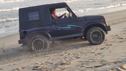 A Man Is Driving A Blue Jeep On A Sandy Beach Near The Ocean GIF