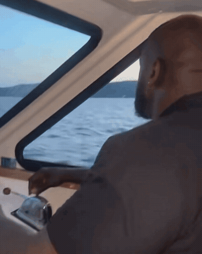 A Man Is Driving A Boat In The Water And Looking Out The Window GIF