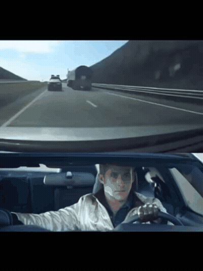 A Man Is Driving A Car On A Highway With A Truck Behind Him GIF