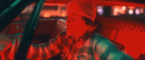 A Man Is Driving A Car With A Beanie On And A Plaid Shirt . GIF