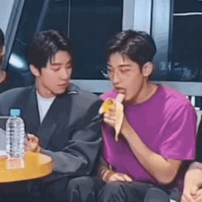 A Man Is Eating A Banana In Front Of A Microphone While Sitting Next To Another Man . GIF