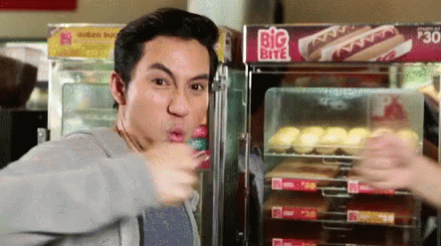 A Man Is Eating A Hot Dog In Front Of A Big Bite Sign GIF