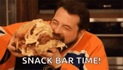 A Man Is Eating A Very Large Sandwich And Saying Snack Bar Time . GIF
