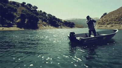 A Man Is Fishing In A Boat On A Lake With Mountains In The Background . GIF