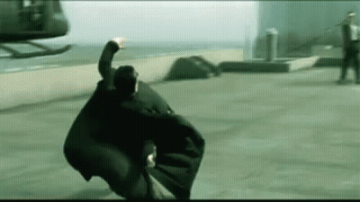 A Man Is Flying Through The Air In Front Of A Helicopter While Wearing A Black Cape . GIF