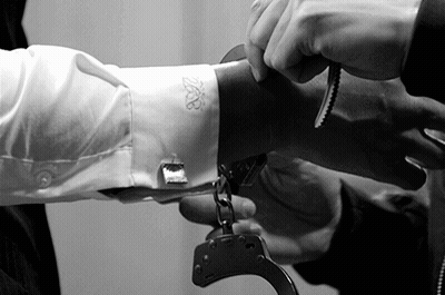 A Man Is Handcuffed To Another Man 'S Arm With A White Shirt With The Letter B On The Sleeve GIF