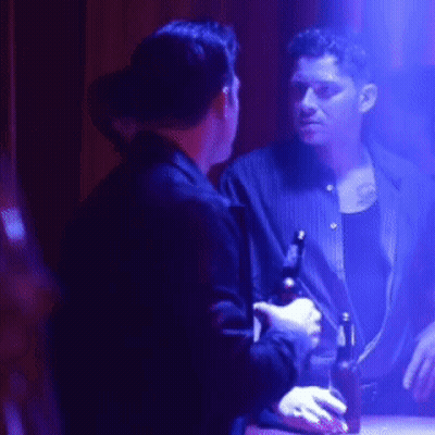 A Man Is Holding A Bottle Of Beer And Talking To Another Man In A Dark Room GIF
