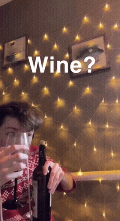 A Man Is Holding A Bottle Of Wine And A Glass In Front Of A String Of Lights And The Word Wine Is On The Wall GIF