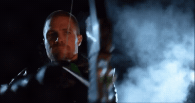 A Man Is Holding A Bow And Arrow In Front Of A Smokey Background . GIF