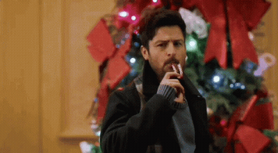 A Man Is Holding A Candy Cane In His Mouth While Standing In Front Of A Christmas Tree . GIF
