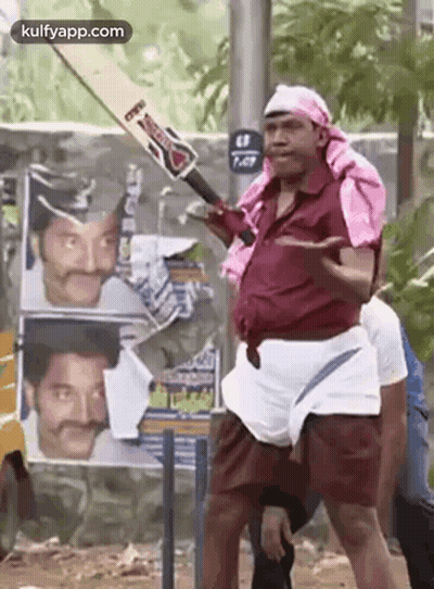 A Man Is Holding A Cricket Bat In Front Of A Wall With Posters Of Actors On It . GIF