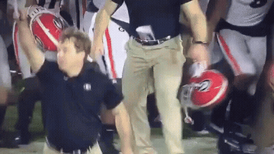 A Man Is Holding A Football In His Hand While A Group Of Football Players Are Standing Behind Him Holding Helmets . GIF