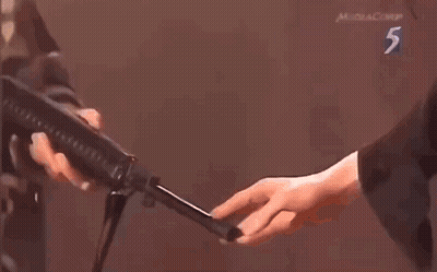 A Man Is Holding A Gun In Front Of A Man Standing Next To A Man Holding A Gun . GIF