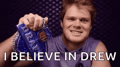 A Man Is Holding A Medal In His Hand And Smiling With The Words `` I Believe In Drew '' Behind Him . GIF