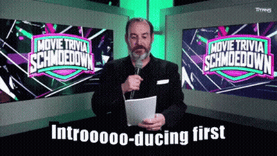 A Man Is Holding A Microphone In Front Of A Screen That Says Movie Trivia Schmoodown GIF