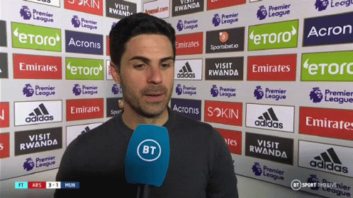 A Man Is Holding A Microphone In Front Of A Wall With Advertisements For Emirates And Etoro GIF
