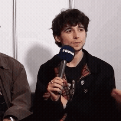 A Man Is Holding A Microphone In His Hand While Sitting In Front Of A Wall . GIF