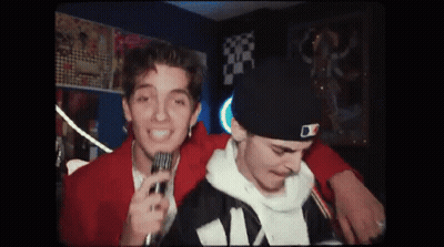 A Man Is Holding A Microphone Next To Another Man In A Hat . GIF