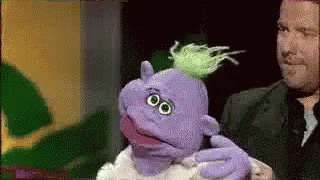 A Man Is Holding A Purple Puppet With A Green Haircut . GIF