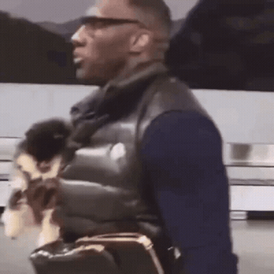 A Man Is Holding A Small Dog In His Arms While Walking Down The Street . GIF