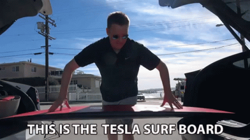 A Man Is Holding A Surfboard In The Back Of A Tesla Car GIF
