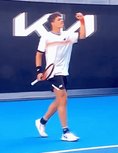A Man Is Holding A Tennis Racquet On A Tennis Court In Front Of A Wall With A Kt Logo GIF