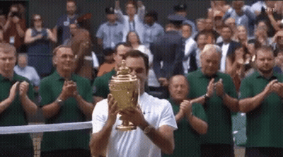 A Man Is Holding A Tennis Trophy In Front Of A Crowd . GIF