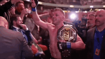 A Man Is Holding A Ufc Heavyweight Championship Belt In A Boxing Ring . GIF