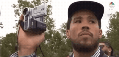 A Man Is Holding A Video Camera In His Hand And Looking At The Camera . GIF