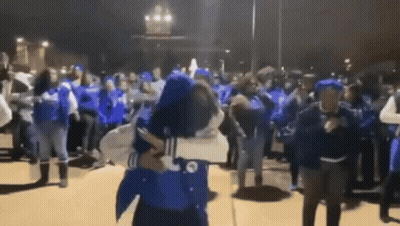 A Man Is Holding A Woman In His Arms In Front Of A Crowd Of People Wearing Blue Shirts . GIF