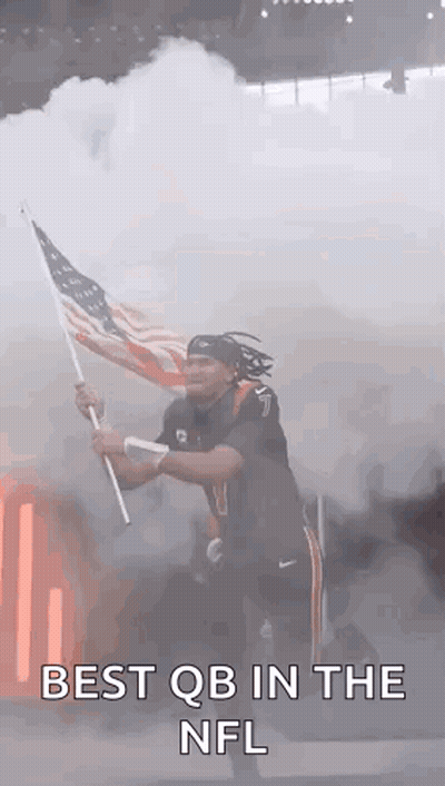 A Man Is Holding An American Flag In Front Of Smoke Coming Out Of A Stadium . GIF
