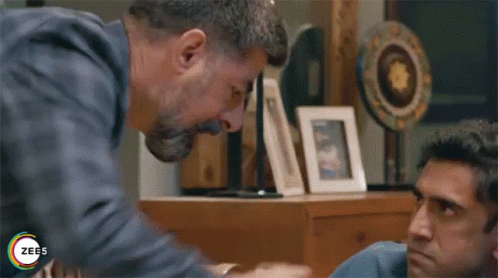 A Man Is Holding Another Man 'S Hand While Sitting At A Table In A Living Room . GIF