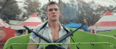A Man Is Holding Two Guns On His Shoulders While Riding A Green Ferris Wheel At A Carnival . GIF