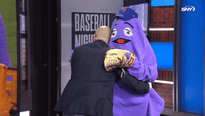A Man Is Hugging A Purple Mascot In Front Of A Baseball Night Sign GIF