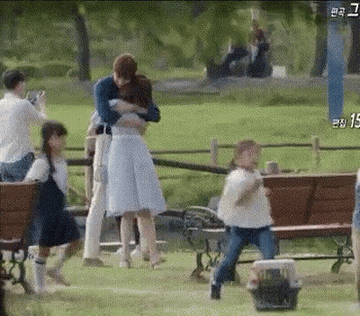 A Man Is Hugging A Woman In A Park While A Group Of People Are Standing Around A Bench . GIF
