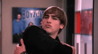 A Man Is Hugging A Woman In A Room With A Poster On The Wall That Says Boyzoo . GIF