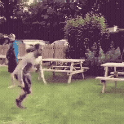 A Man Is Jumping In The Air In A Field With Picnic Tables In The Background GIF