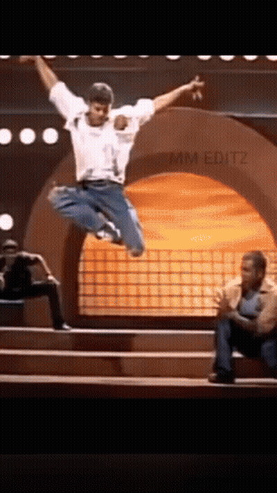 A Man Is Jumping In The Air On A Stage While Another Man Sits On The Steps . GIF