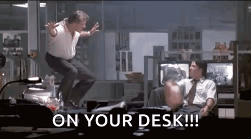 A Man Is Jumping In The Air While Another Man Sits At A Desk In An Office . GIF