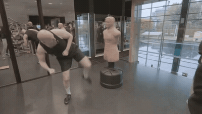 A Man Is Kicking A Punching Bag In A Gym While Another Man Watches GIF