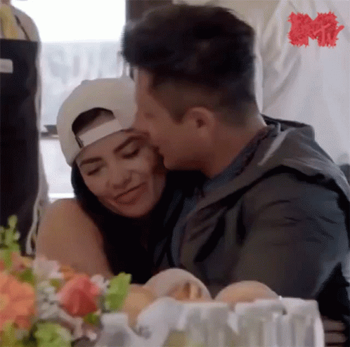 A Man Is Kissing A Woman On The Cheek While Sitting At A Table With Flowers . GIF
