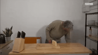 A Man Is Kneeling Down On A Wooden Table With A Knife Block And A Cutting Board . GIF
