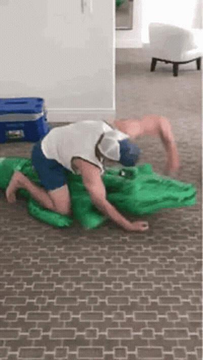 A Man Is Kneeling On The Floor With A Green Inflatable Alligator . GIF