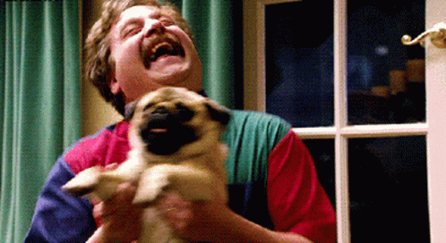 A Man Is Laughing While Holding A Small Pug Dog GIF