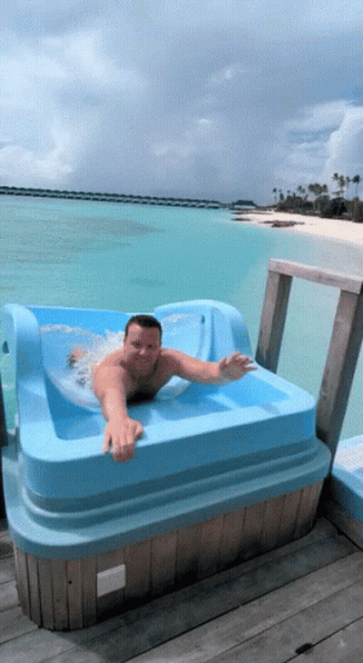 A Man Is Laying In A Blue Tub On A Dock Overlooking The Ocean GIF