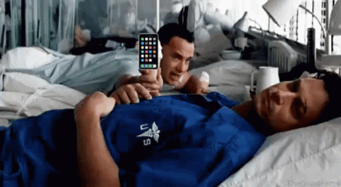 A Man Is Laying In A Hospital Bed While Another Man Is Holding A Cell Phone GIF
