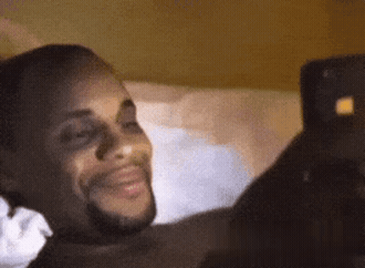 A Man Is Laying On A Bed Looking At A Cell Phone And Smiling . GIF