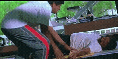 A Man Is Laying On A Bed While Another Man Is Standing Next To Him . GIF